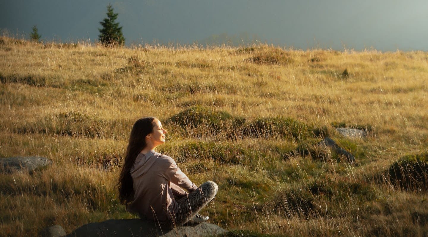 Woman sitting in a field of grass with her eyes closed listening to the calm breeze.