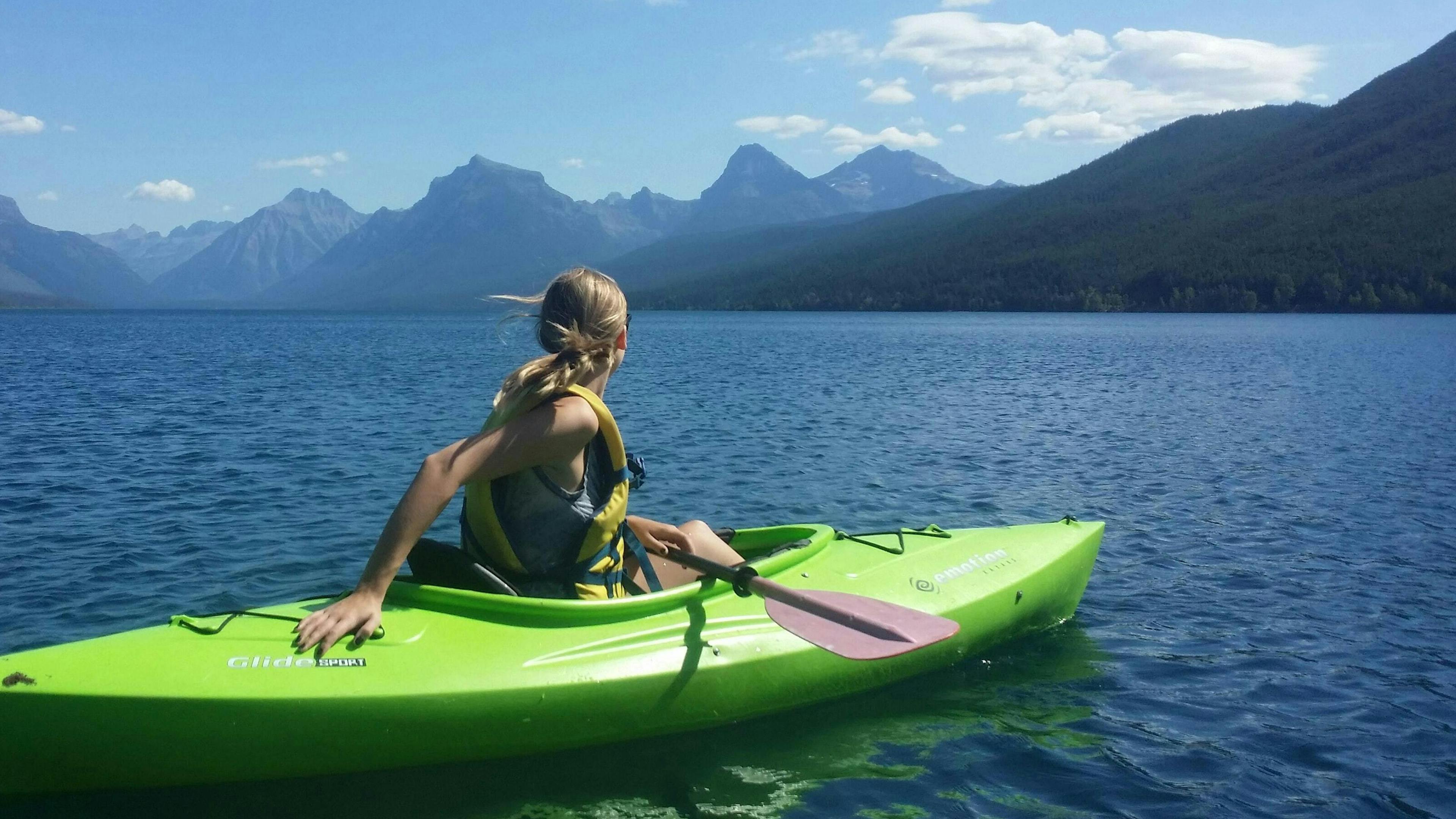 A woman on the water in a green kayak looking towards a vast mountain range.
