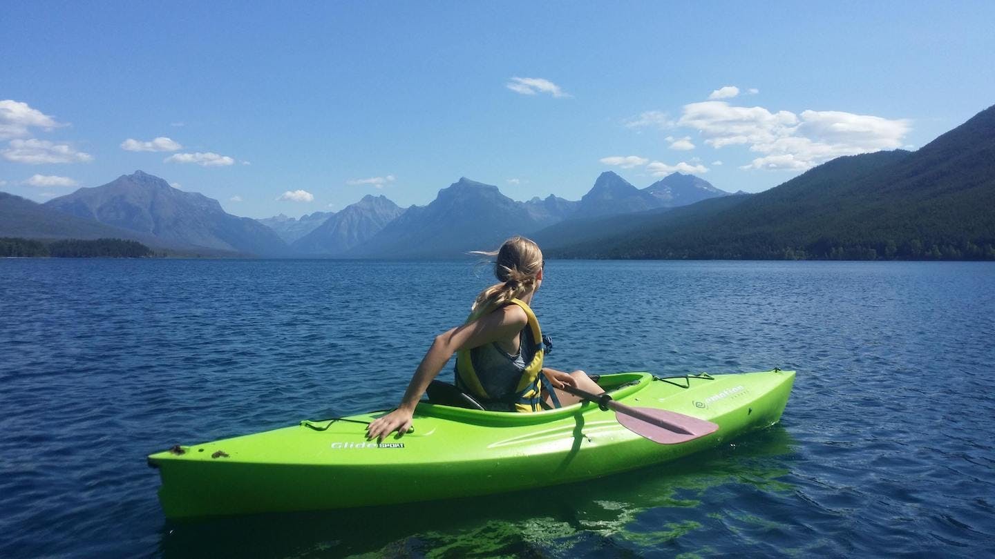 A woman on the water in a green kayak looking towards a vast mountain range.